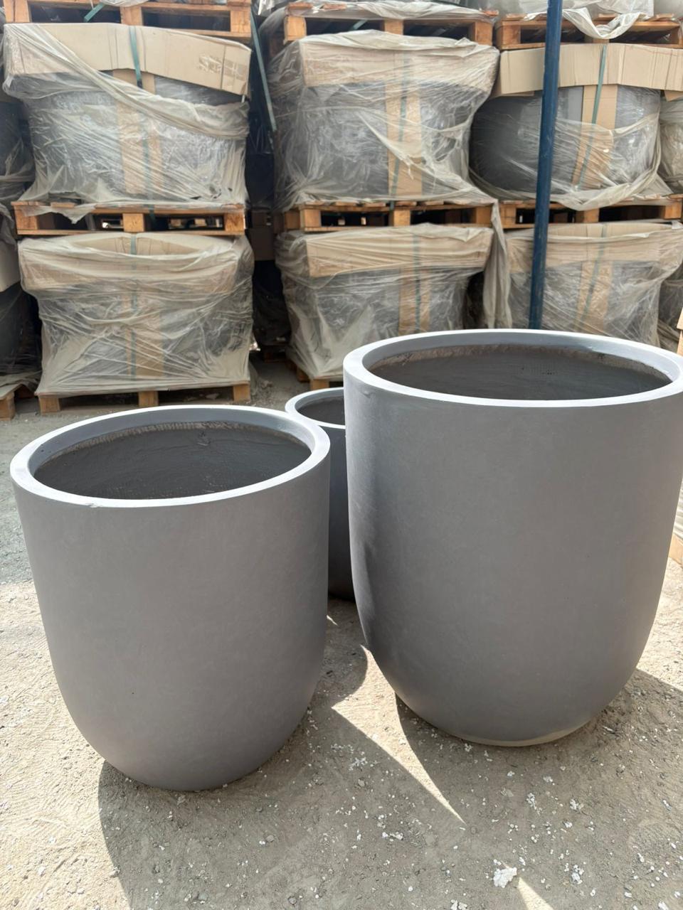Outdoor and Indoor Large Pot Plant UAE. Two large gray planters on a concrete surface with pallets and wrapped items in the background.