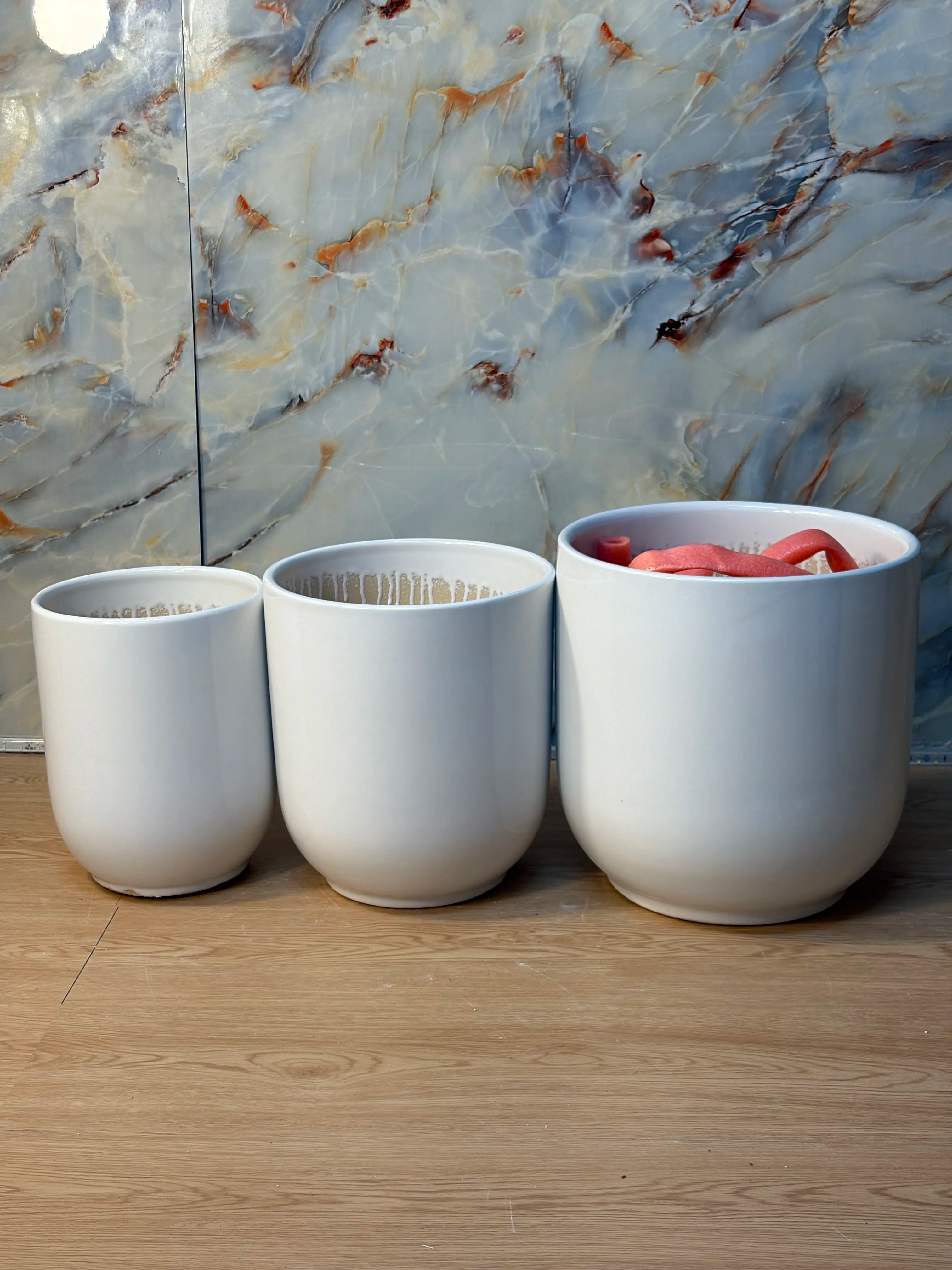 White Glossy Ceramic Plants Pot UAE. Three white ceramic planters on a wooden floor with a marble wall in the background